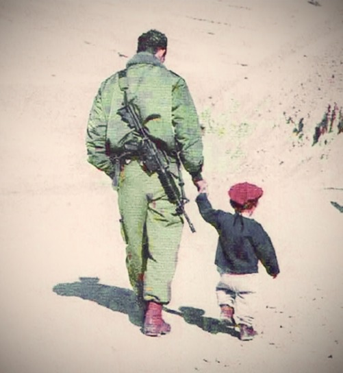 An Israeli paratrooper walking away from the camera, holding the hand of a small child in a red hat.